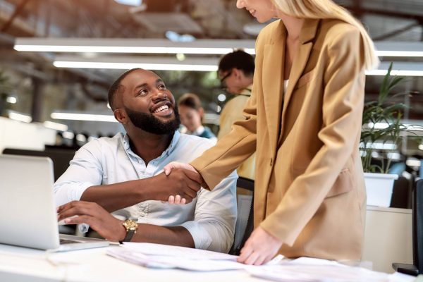 Young cheerful African American cheerful man and beautiful business woman shaking hands while working together in the modern office.
